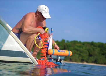 A middle aged man wearing a white cap and swimming shorts is squatting on the rear yacht duckboard placing a DiveBuddy portable dive hookah compressor into the water with his right hand while holding the bright orange coiled hookah breathing and dive hookah regulator. Tropical island beach and trees in the background Picture